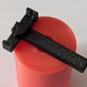 Puede incluir: Un objeto negro en forma de T, texturizado, descansa sobre una bobina cilíndrica roja. El objeto parece estar impreso en 3D. La bobina tiene una superficie texturizada y es de color rojo vibrante. El fondo es blanco liso.