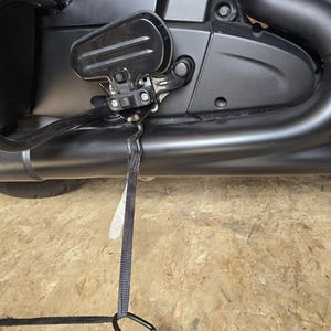 May include: Close-up of a black motorcycle engine and exhaust system. The image shows the engine's side, with a black exhaust pipe and a black strap hanging down. The motorcycle parts are mounted on a wooden surface.