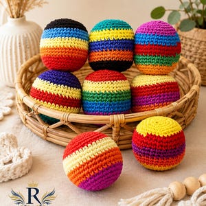 Puede incluir: Una colección de bolas de ganchillo coloridas en una cesta de mimbre. Cada bola presenta rayas horizontales de varios colores, incluyendo rojo, naranja, amarillo, verde, azul y morado. Las bolas miden aproximadamente 7.6 cm de diámetro y están dispuestas sobre una superficie de tono neutro.