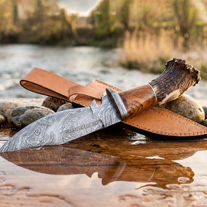 Puede incluir: Un cuchillo de caza con una hoja de acero estampada, mango de madera y pomo de asta descansa en una funda de cuero marrón. El cuchillo se exhibe sobre una superficie de madera con un reflejo de agua.