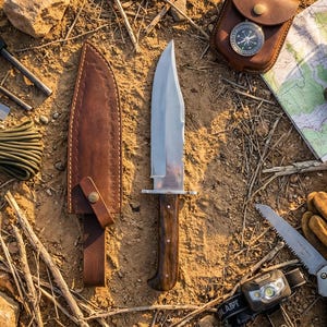Peut inclure: Un grand couteau de chasse en acier inoxydable avec un manche en bois et un étui en cuir marron. Le couteau est entouré d'autres équipements de plein air, dont une boussole, une carte et une lampe frontale.