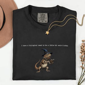 Puede incluir: Camiseta negra Comfort Colors con un diseño de lagarto con sombrero de mago y libro. El texto dice: "I have a biological need to be a little bit weird today." Un collar con colgante de estrella dorada está sobre la camiseta. Talla XL.