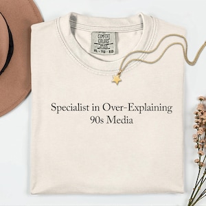 May include: Cream-colored t-shirt with the text "Specialist in Over-Explaining 90s Media" in black. A gold star pendant necklace and a brown hat are also visible. The shirt has a Comfort Colors tag.