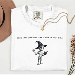 Könnte beinhalten: Weißes Comfort Colors T-Shirt mit dem Text "I have a biological need to be a little bit weird today." Auf der Vorderseite ist ein Cartoon-Alien mit Hexenhut und Buch abgebildet. Eine goldene Stern-Anhänger-Halskette liegt auf dem Shirt.