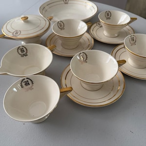 May include: A cream-colored porcelain tea set featuring a gold-rimmed design and a central letter B emblem. The set includes a lidded sugar bowl, a creamer, a shallow bowl, and four teacups with matching saucers, all decorated with gold accents.