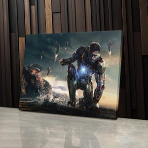 Op de afbeelding: Canvas print met een knielende figuur in een metallic en rood pantserpak, met een gloeiend borststuk. De achtergrond toont een dramatische lucht, explosies en figuren in vlucht, wat een superheldenthema oproept.