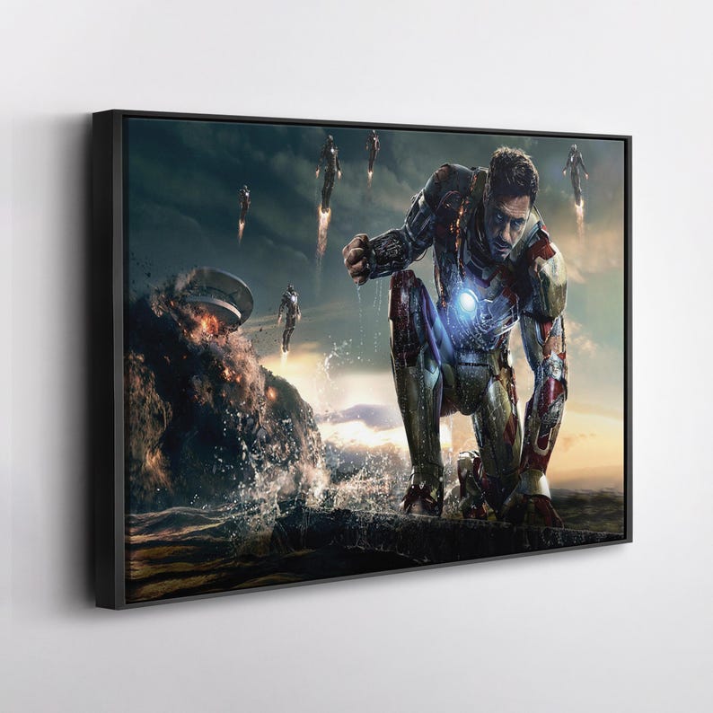 Op de afbeelding: Ingelijste kunstwerk met een knielende figuur in een metalen pak met een gloeiend borststuk, tegen een dramatische oceaan- en hemelachtergrond. Er zijn verschillende vliegende figuren zichtbaar, met een explosie in de verte.