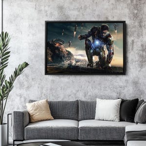 Op de afbeelding: Een ingelijste canvas print met Iron Man die knielt op een rotsachtige oever, met andere figuren die op de achtergrond vliegen. Het kunstwerk heeft een filmische uitstraling, met de nadruk op het pantser van het personage en de omgeving.