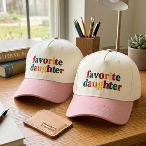 May include: Two baseball caps with a white crown and a pink brim. The phrase "favorite daughter" is embroidered in colorful letters on the front. A small leather cardholder with the text "best daughter in the world" is also visible.