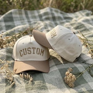May include: Two baseball caps with beige crowns and brown visors. One cap has the word "CUSTOM" embroidered in a matching beige thread. The other cap has the word "TOM" embroidered in a matching beige thread. The caps are resting on a green and white checkered blanket.