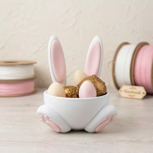Puede incluir: Una cesta de Pascua de cerámica blanca con forma de conejito, con detalles en rosa, llena de huevos de colores pastel y bombones envueltos. La cesta presenta orejas y patas de conejo. Rollos de cinta en el fondo.