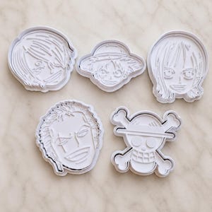 以下が含まれることがあります： キャラクターの顔とドクロのエンブレムをかたどった、5種類の白いプラスチック製クッキー型セット。詳細な顔のパーツ、髪型、麦わら帽子が、成形されたプラスチック構造で表現されています。