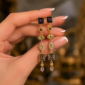 May include: A pair of gold-tone drop earrings featuring a vertical arrangement of multi-colored gemstones. Each earring includes a square purple stone, a small black accent, a light green oval, a yellow marquise, a smoky gray rectangle, and a teardrop.
