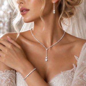 Può includere: Un set di gioielli composto da una collana a Y, un bracciale abbinato e un orecchino pendente, tutti caratterizzati da pietre trasparenti a taglio rotondo incastonate in metallo argentato. La collana presenta un pendente verticale centrale e il bracciale è sottile.