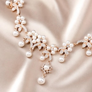 May include: A gold-toned necklace featuring a floral-inspired design with white faux pearls and clear crystals. The piece consists of interconnected metal segments shaped like leaves and flowers, with dangling pearl accents throughout the structure.