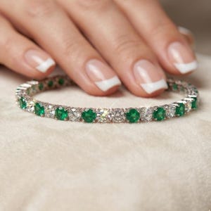 Op de afbeelding: Een zilveren armband met afwisselend ronde smaragd- en diamant edelstenen. De armband ligt op een witte ondergrond. De edelstenen zijn helder en sprankelend.