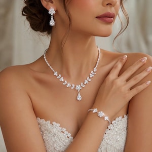 Könnte beinhalten: Ein silberfarbenes Schmuckset bestehend aus einer Halskette, einem Armband und Ohrhängern. Jedes Stück ist mit klaren, facettierten Steinen in Blumenmustern verziert, wobei die Halskette einen zentralen tropfenförmigen Steinanhänger besitzt.