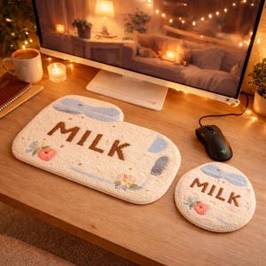 May include: A beige mousepad and coaster set with the word "MILK" in brown, and floral and blue accents. The mousepad has a unique shape, and the coaster is round. The set is on a wooden desk.
