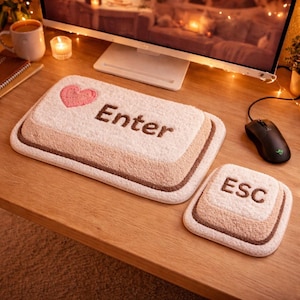 Puede incluir: Un juego de alfombras beige y marrón con forma de teclas de ordenador. La alfombra más grande tiene la palabra "Enter" y un corazón rosa. La alfombra más pequeña tiene la palabra "ESC". Las alfombras están sobre una superficie de madera.