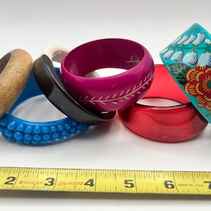May include: A collection of six assorted bangles including a natural wood bracelet, a blue textured bangle, a black and white bangle, a magenta bangle with etched leaf patterns, a translucent red bangle, and a turquoise bangle with floral designs.