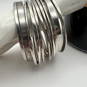 May include: A set of multiple silver-toned metal bangle bracelets with a smooth, polished finish. The bangles vary slightly in width and thickness, featuring a reflective surface that shows subtle signs of wear and oxidation on some bands.
