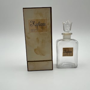 May include: A clear glass perfume bottle with a tulip-shaped stopper featuring the letter R, alongside a rectangular cardboard box. Both the bottle and the box display a tan label with the text Réplique and Raphael Paris in black script.