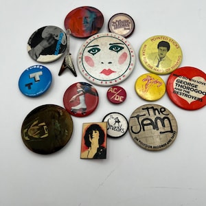 May include: A collection of circular and rectangular pinback badges featuring various music-themed graphics, including band names like The Jam, AC/DC, and The Tubes, portraits of musicians, a guitar-shaped pin, and text such as George Thorogood.