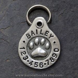 May include: Silver dog tag with a paw print and the name "BAILEY" engraved on it. The tag also has numbers 1, 2, 3, 4, 5, 6, 7, 8, 9, and 0 engraved around the paw print.