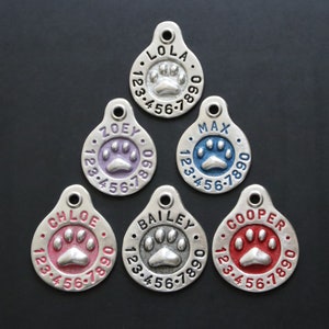 May include: Six silver dog tags with engraved names and paw prints. The names are Lola, Zoey, Max, Chloe, Bailey, and Cooper. Each tag has a different colored enamel paw print: pink, purple, blue, red, gray, and red.