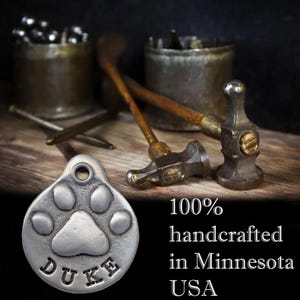 May include: A silver dog tag featuring a paw print and the name "DUKE" engraved on it. The tag is handcrafted in Minnesota, USA. The image shows the tag alongside tools used in the crafting process.