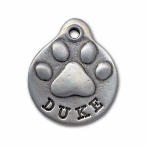 May include: Silver dog tag featuring a paw print design with the name "DUKE" engraved on it.