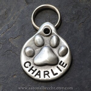 May include: Silver dog tag featuring a paw print design with the name "CHARLIE" engraved on it.