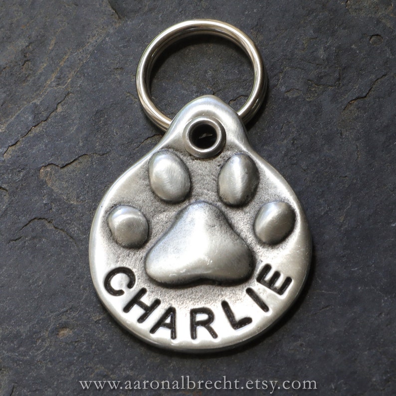 May include: Silver dog tag featuring a paw print design with the name "CHARLIE" engraved on it.