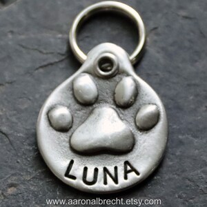 May include: Silver dog tag featuring a paw print design with the name "LUNA" engraved on it. The tag has a ring for attaching to a collar.