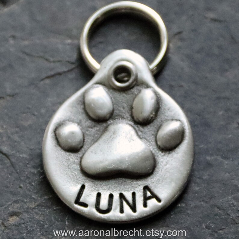 May include: Silver dog tag featuring a paw print design with the name "LUNA" engraved on it. The tag has a ring for attaching to a collar.