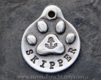 Puede incluir: Chapa de perro de plata con un diseño de huella de pata y el nombre "SKIPPER" grabado en el borde. Un ancla está grabada dentro de la huella de pata.