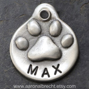 May include: Silver dog tag featuring a paw print design with the name "MAX" engraved on it.