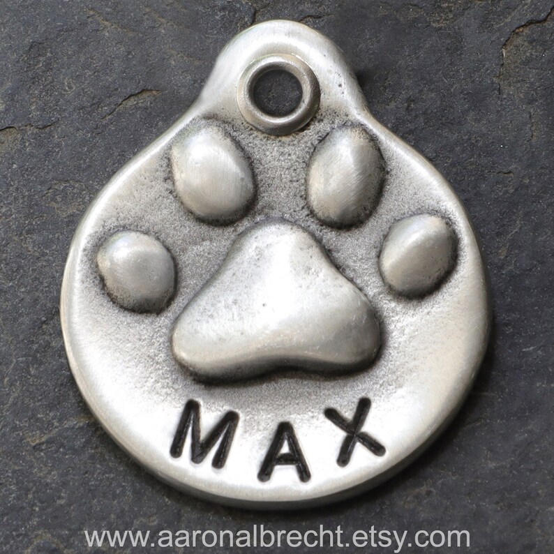 May include: Silver dog tag featuring a paw print design with the name "MAX" engraved on it.
