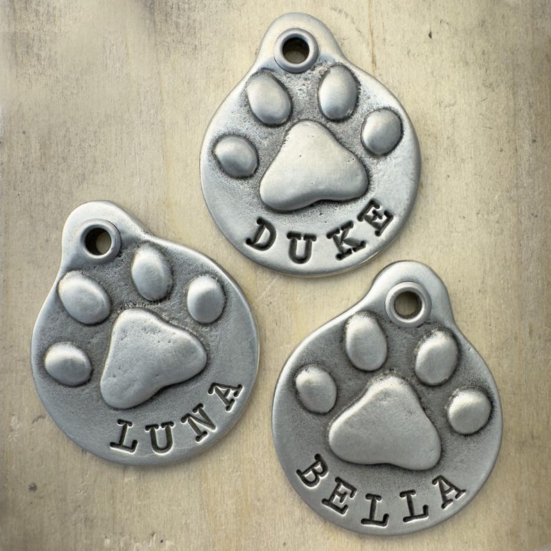 May include: Three silver dog tags featuring a paw print design. The tags are engraved with the names "Duke", "Luna", and "Bella".
