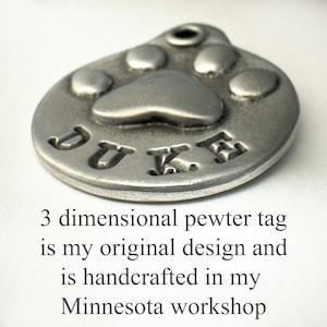 May include: A silver pewter dog tag featuring a paw print design and the name "DUKE" engraved on it. The tag is 3-dimensional and handcrafted.