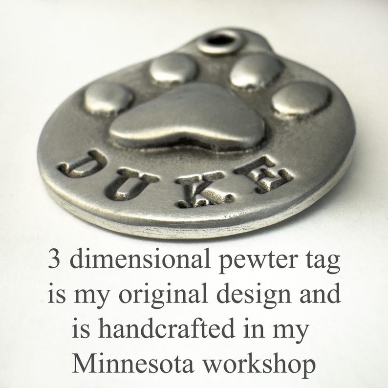 May include: A silver pewter dog tag featuring a paw print design and the name "DUKE" engraved on it. The tag is 3-dimensional and handcrafted.