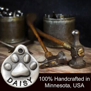 May include: A silver pet tag featuring a paw print design with the name "DAISY" engraved on it. The tag is handcrafted in Minnesota, USA.