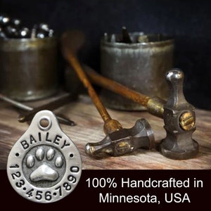 May include: Silver dog tag with the name "BAILEY" and a paw print design. The tag is surrounded by numbers 123.456.7890. The tag is sitting on a wooden surface with two metal hammers.