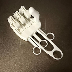Puede incluir: Un modelo de mano articulada blanca impreso en 3D, con palma, dedos y muñeca. Los dedos están separados y parecen ser posables. El modelo está sobre una superficie oscura y texturizada.
