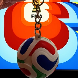May include: A spherical keychain featuring a white base with swirling, raised patterns in red, blue, and green. The ball is attached to a silver-toned metal split ring by a short chain. The design mimics the appearance of a soccer ball.