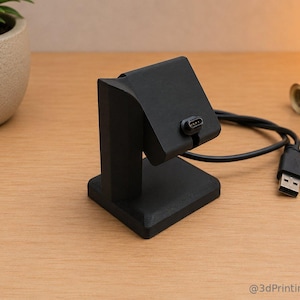 Op de afbeelding: Een zwarte oplaadstandaard met een USB-kabel. De standaard heeft een vierkante basis en een verticale steun met een gebogen bovenkant. Een kleine plant in een pot is zichtbaar op de achtergrond.