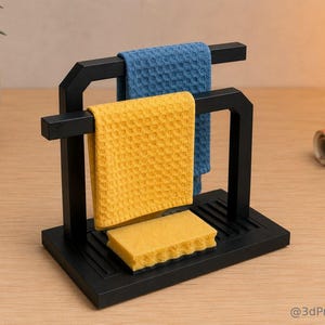 Könnte beinhalten: Ein schwarzer, geometrischer Küchenhandtuch- und Schwammhalter mit einem gelben Schwamm und zwei Waffelhandtüchern, eines blau und eines gelb. Der Halter steht auf einer Holzoberfläche.