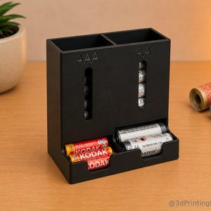 Könnte beinhalten: Ein schwarzer Batterie-Organizer aus Kunststoff mit zwei vertikalen Fächern, beschriftet mit AAA und AA, mit Sichtfenstern für die Batterien und einem unteren Fach für lose rote Kodak- und silberne AmazonBasics-Batterien.