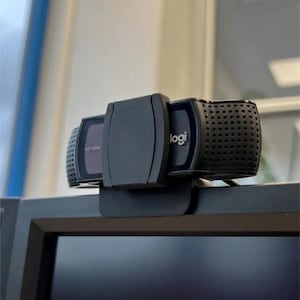 May include: A black Logitech webcam with the logo visible. The webcam is mounted on top of a computer monitor. The webcam has a rectangular design with a central lens and two side sections with a perforated design.
