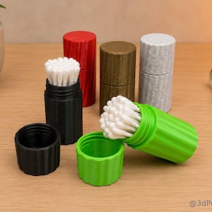 Può includere: Una collezione di contenitori cilindrici stampati in 3D in vari colori, tra cui nero, rosso, verde e grigio. Alcuni contenitori sono aperti, rivelando bastoncini di cotone bianchi. I contenitori sono su una superficie di legno.